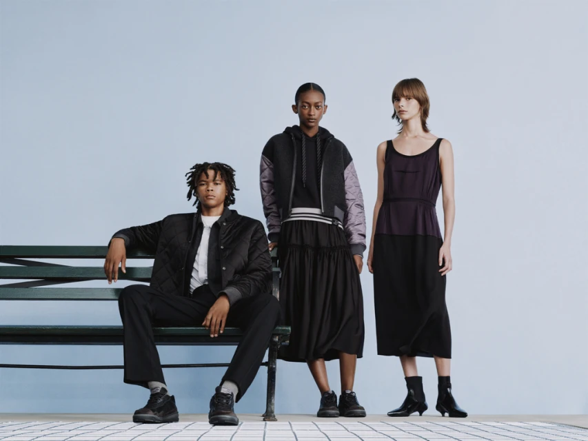 Three fashion models pose in black and grey clothing on a bench against a light blue backdrop in a studio setting.