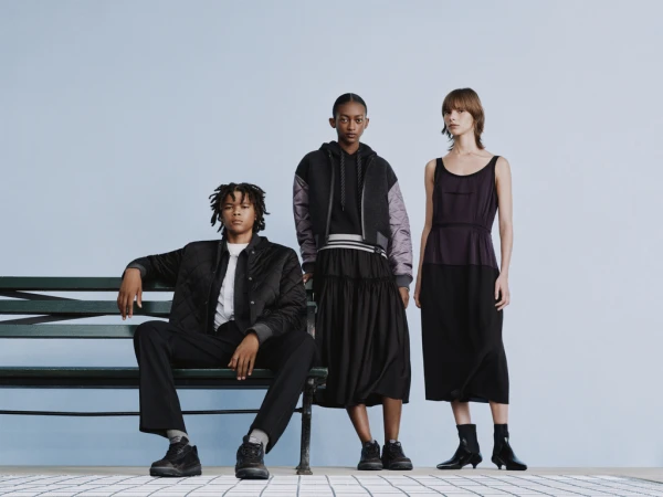 Three fashion models pose in black and grey clothing on a bench against a light blue backdrop in a studio setting.