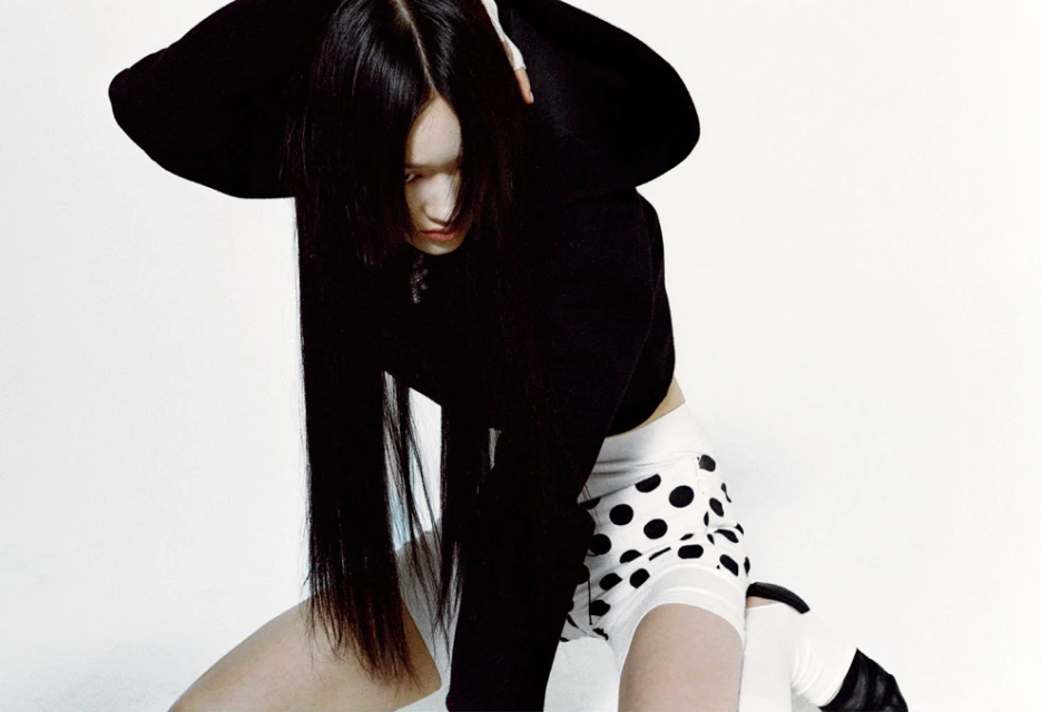 Woman with long black hair poses in a black crop top and white polka dot shorts against a stark white background in a fashion shot.