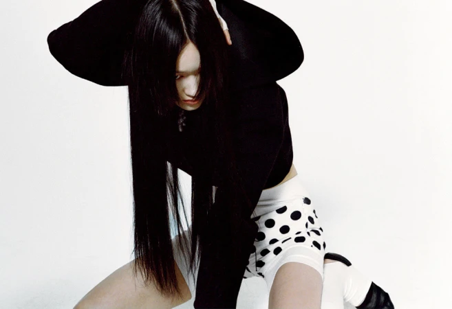 Woman with long black hair poses in a black crop top and white polka dot shorts against a stark white background in a fashion shot.