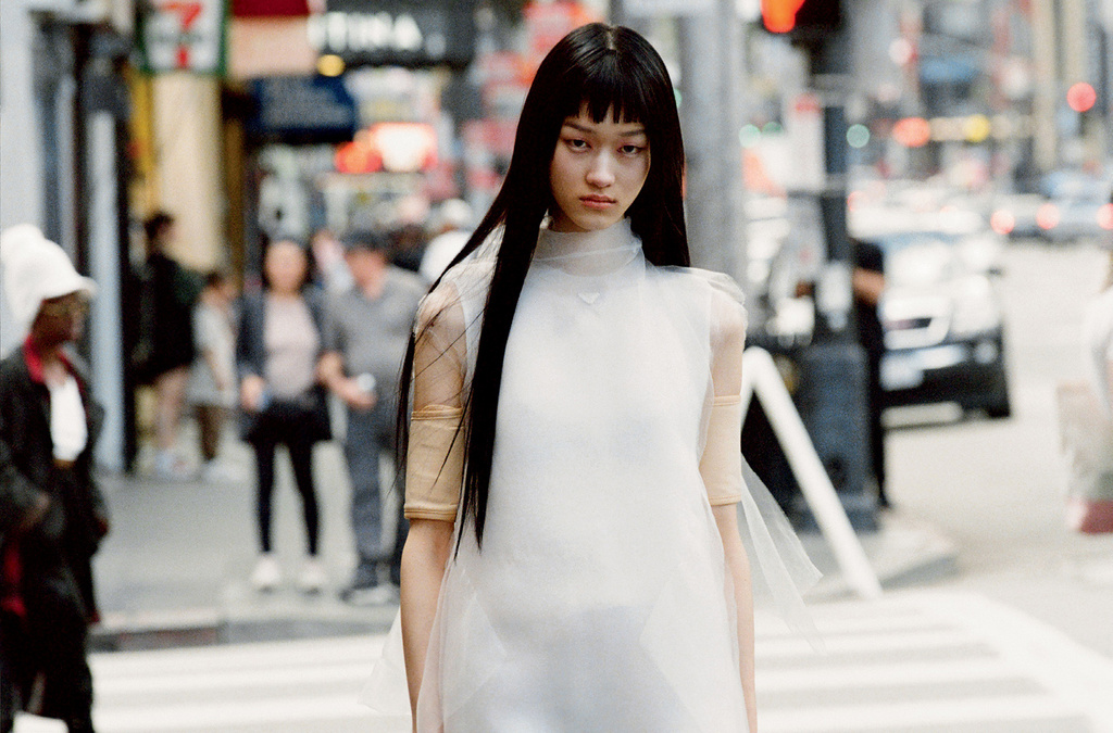Model with long black hair wearing a sheer white sleeveless top walking on a city street with blurred background