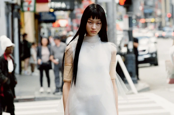 Model with long black hair wearing a sheer white sleeveless top walking on a city street with blurred background