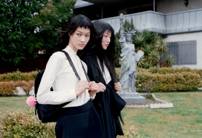 Two Asian women in white shirts and black skirts stand outdoors near a statue of liberty replica, showcasing a blend of fashion and environment.