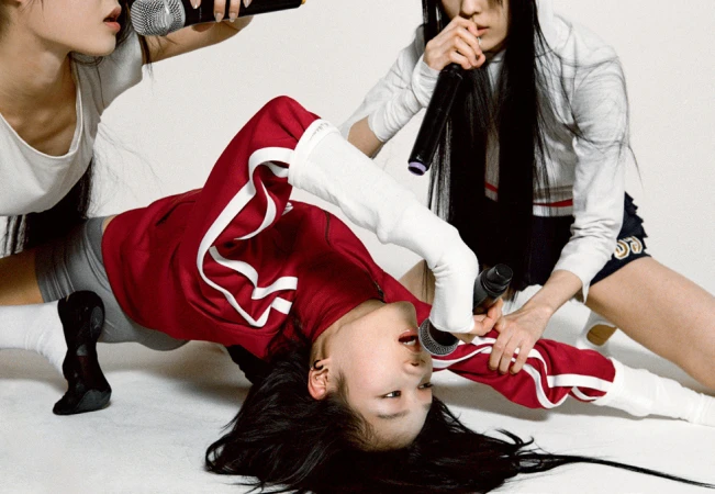 Three women perform in a studio, singing into microphones. One woman is on the floor doing a split while holding a microphone.
