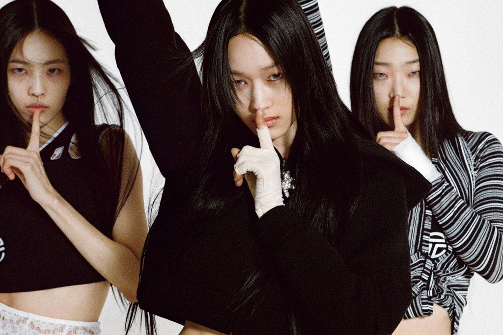 Three Asian women in a studio setting with finger to lips in a 'be quiet' gesture, wearing dark clothing against a white backdrop.