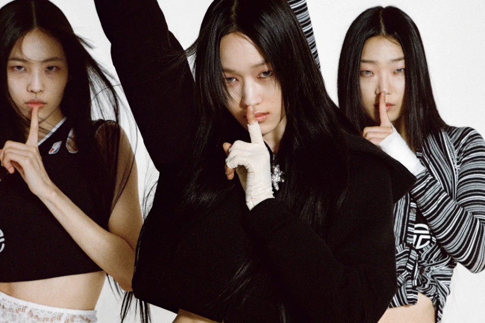 Three Asian women in a studio setting with finger to lips in a 'be quiet' gesture, wearing dark clothing against a white backdrop.