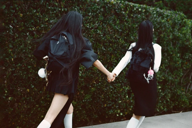 Two young women with long black hair holding hands and walking along a green hedge.