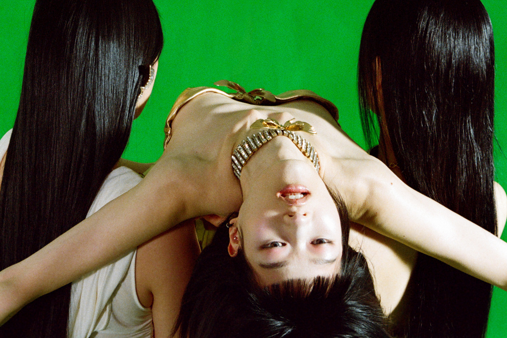 Three women are captured in a studio shot against a green backdrop. One woman is centered, wearing gold jewelry. All have sleek, dark hair.