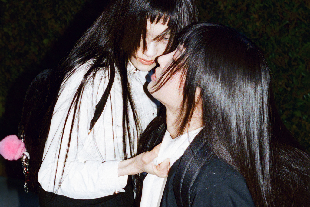 Two women with long black hair and bangs in a close moment, one in a white shirt and the other in a black jacket.