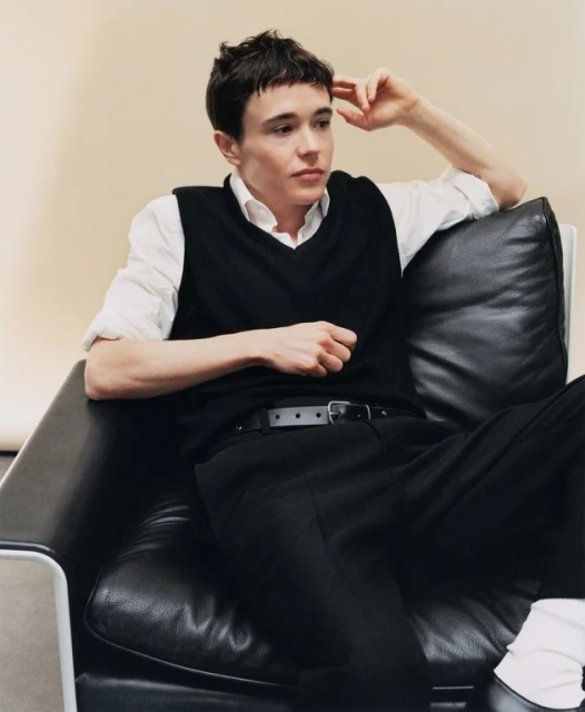 Young adult sitting in a black leather armchair wearing a white shirt, black vest and black pants in an indoor environment.