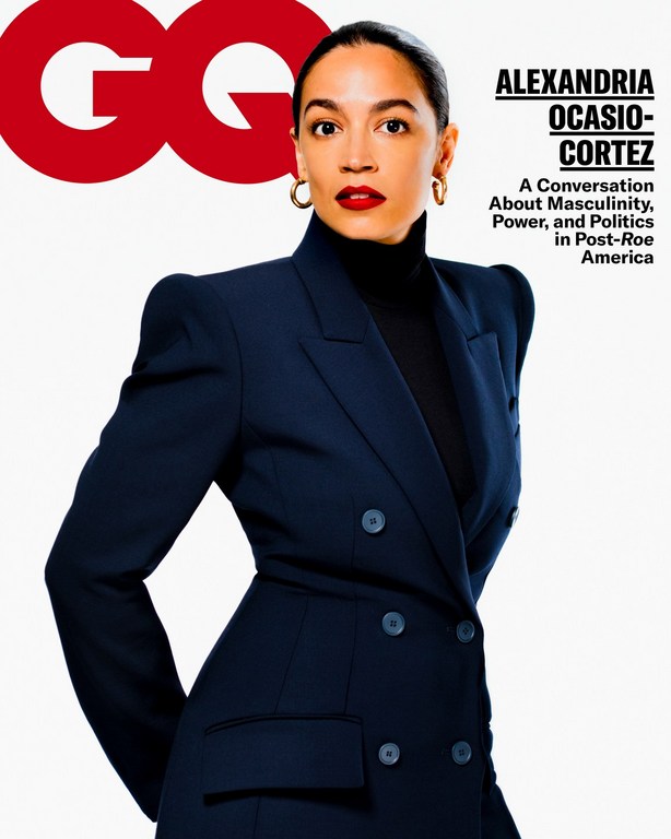 Alexandria Ocasio-Cortez poses on the cover of GQ magazine wearing a navy blue suit with red lipstick and gold hoop earrings.