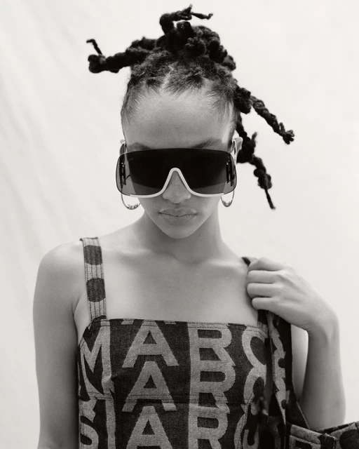 Woman with locs wears sunglasses and a top with 'MARC JACOBS' lettering in a black and white portrait against a light backdrop.
