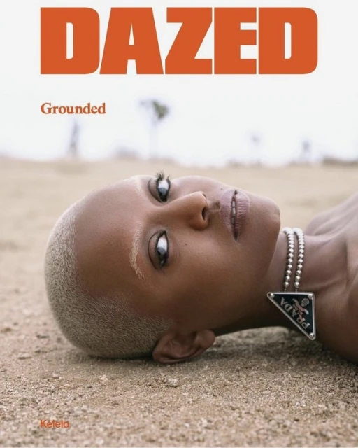 Close-up of a black woman with a shaved head lying on sand, wearing a Prada necklace for a Dazed magazine cover with orange text.
