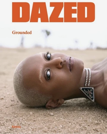 Close-up of a black woman with a shaved head lying on sand, wearing a Prada necklace for a Dazed magazine cover with orange text.