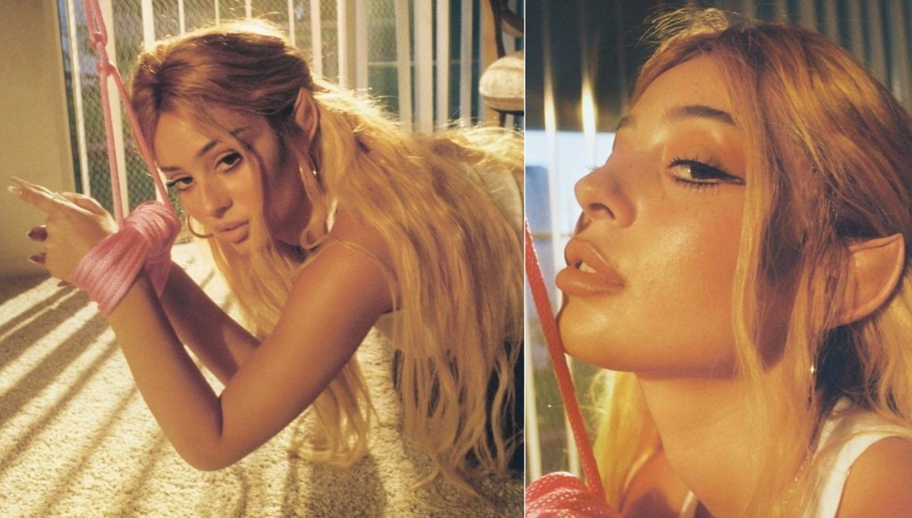 Portrait of a woman with long blonde hair and pointed ears, hands wrapped with pink rope, kneeling on all fours inside.