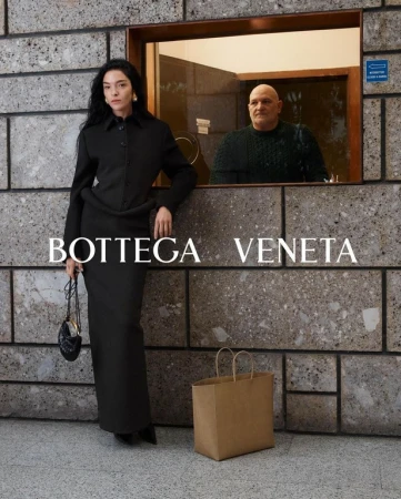 Woman poses in Bottega Veneta outfit with purse and shopping bag, alongside a man peering out a window, in an urban setting.