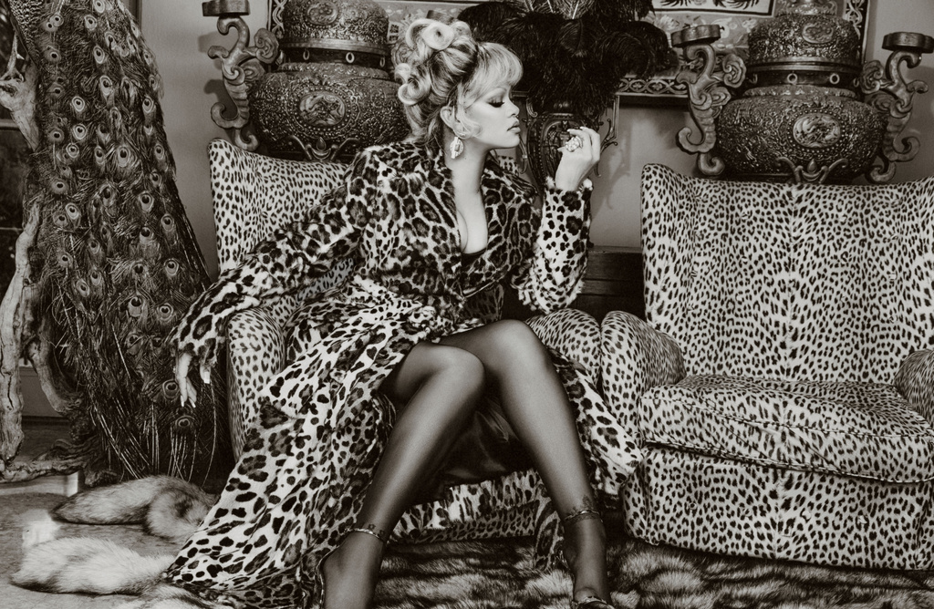 Woman in a leopard print coat sits in a matching chair in a monochrome portrait, surrounded by peacock feathers and ornate objects.