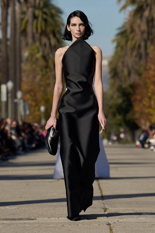 Runway fashion model in black evening gown holding a clutch, walking outdoors during daytime, crowd of spectators in the background.