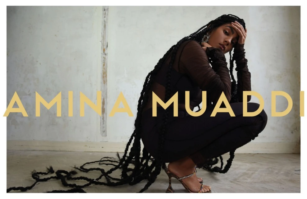Woman posing in a crouching position wearing a dark outfit and heels, adorned with long braids, against a neutral wall with a large text overlay.