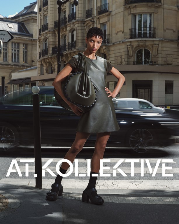 Model standing on a Parisian street holding a large dark leather bag, wearing a sleeveless leather dress and platform shoes.