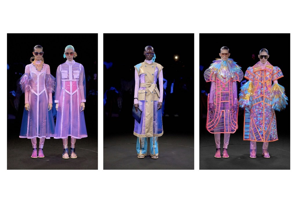 Fashion runway presentation featuring three models wearing uniquely designed transparent coats in a dark indoor setting with blue lighting.