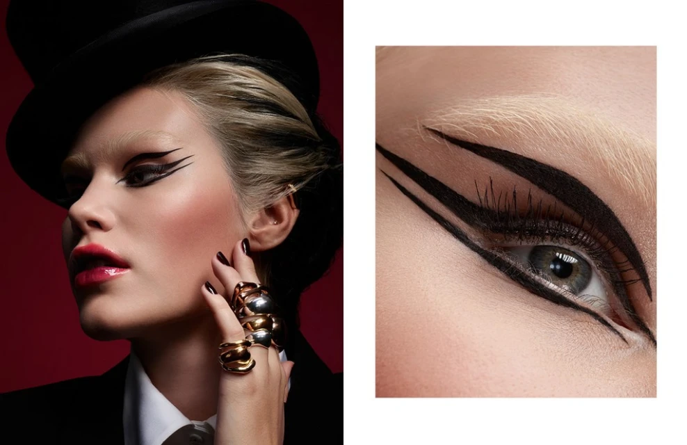 Close-up studio shot of a fair-skinned adult female in a top hat with creative black graphic eye makeup, striking gold rings, and deep red lips.