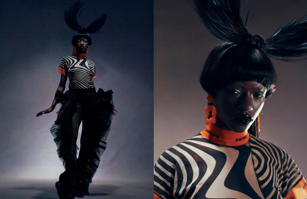 A striking fashion shot features a dark-skinned model with an elaborate updo, bold makeup, and a unique striped outfit against a neutral backdrop.