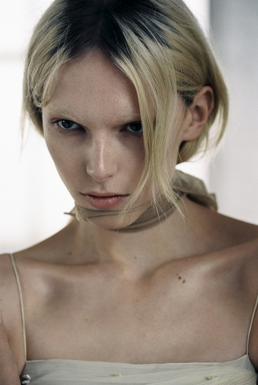 Portrait of a fair-skinned woman with blonde hair and a beige garment, looking at the camera with a serious facial expression indoors.
