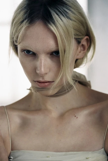 Portrait of a fair-skinned woman with blonde hair and a beige garment, looking at the camera with a serious facial expression indoors.