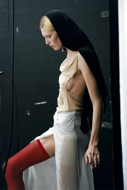 Artistic fashion portrait of a blond woman in a black head covering, beige top, sheer white skirt, and red stockings against a dark backdrop.