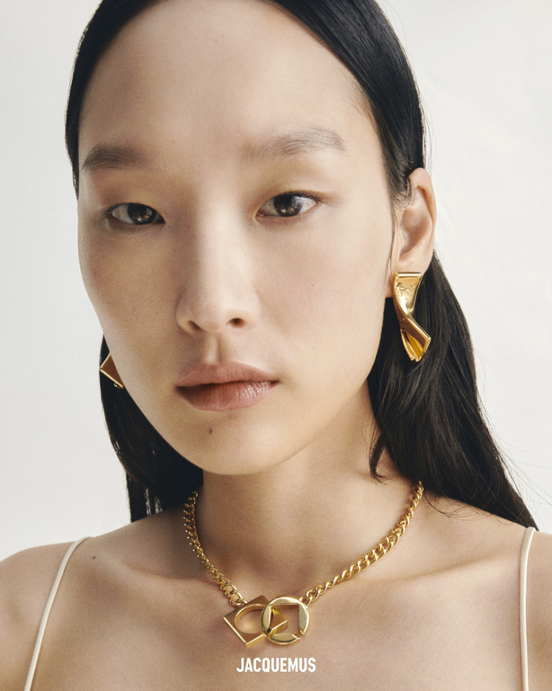 Close-up portrait of an East Asian woman wearing gold jewelry: earrings and a chain necklace with logo charms against a white background.