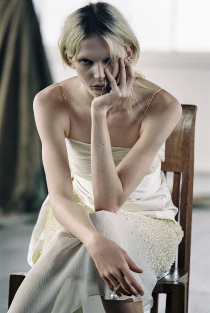 Elegant woman in a light-colored dress sits on a wooden chair with her hand to her face in an indoor environment.
