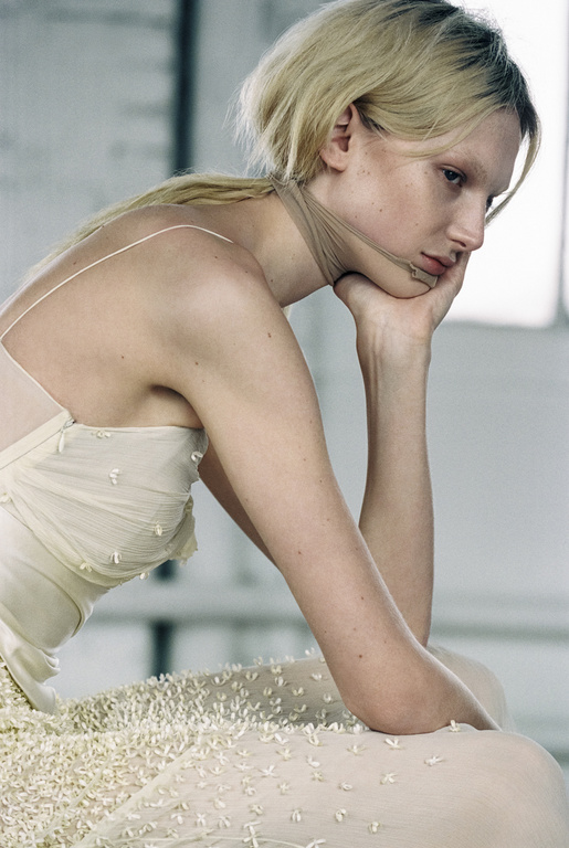 Profile portrait of a blonde woman resting her chin on her hand, wearing a cream textured sleeveless dress.
