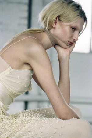 Profile portrait of a blonde woman resting her chin on her hand, wearing a cream textured sleeveless dress.