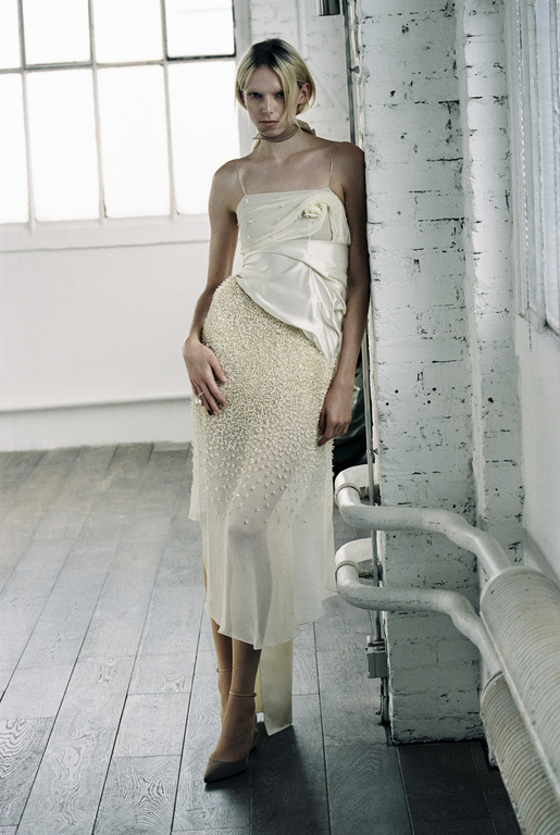 Full-length shot of a slender adult female fashion model in a white gown posing against a brick wall near a window in an indoor space.