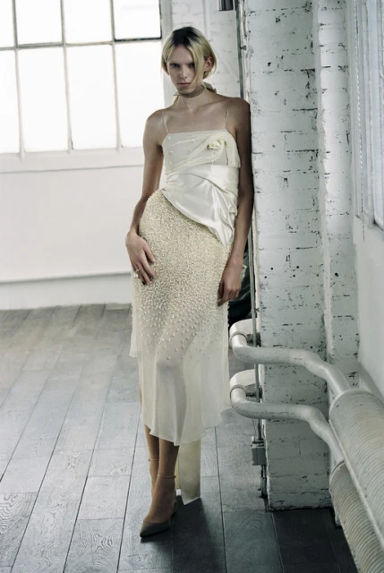 Full-length shot of a slender adult female fashion model in a white gown posing against a brick wall near a window in an indoor space.