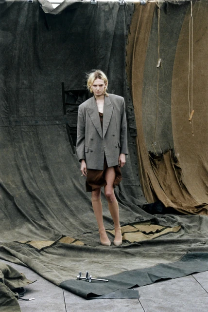 A fashionable person with blond hair poses indoors wearing an oversized grey blazer, brown dress, and tan heels in front of draped fabric.