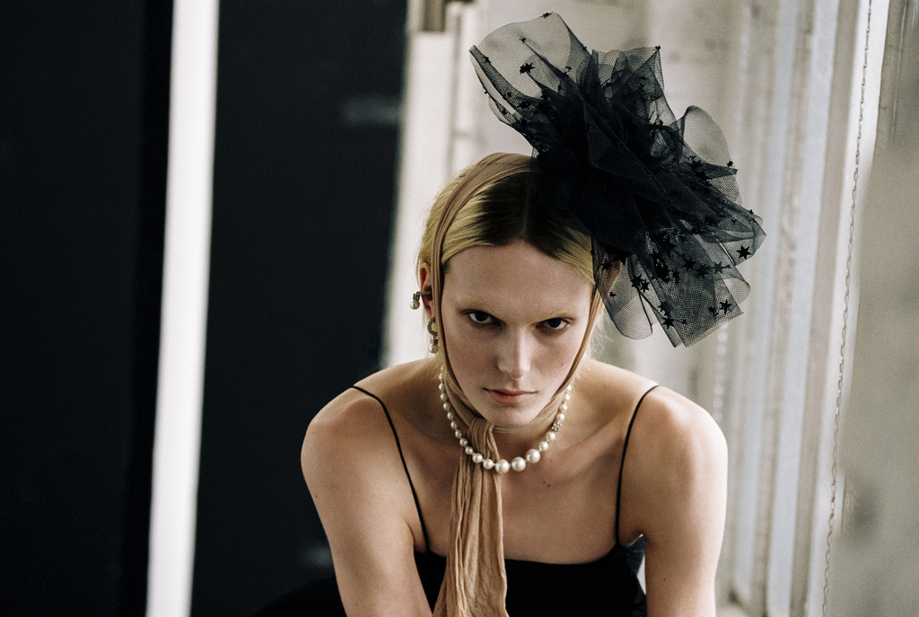 Striking studio portrait of a woman with blonde hair, adorned with a black star-studded headpiece and pearl necklace, gazing directly at the viewer.