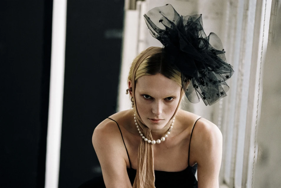 Striking studio portrait of a woman with blonde hair, adorned with a black star-studded headpiece and pearl necklace, gazing directly at the viewer.