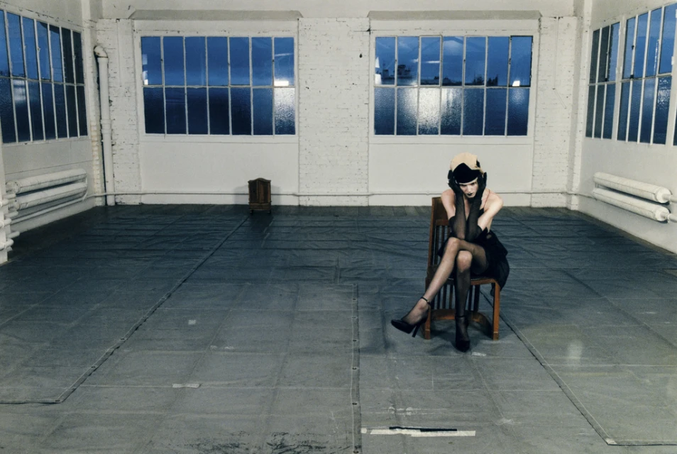 Adult woman in a black dress sits in a wooden chair in an empty studio with large windows and white brick walls, wearing a hat and heels.