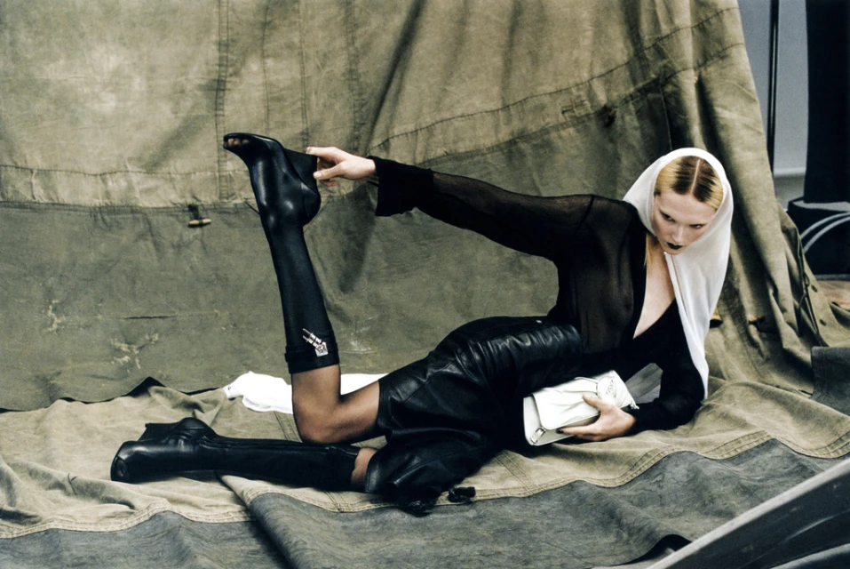 Fashionable woman in leather skirt and boots poses with purse in front of canvas tent for an editorial fashion shot.