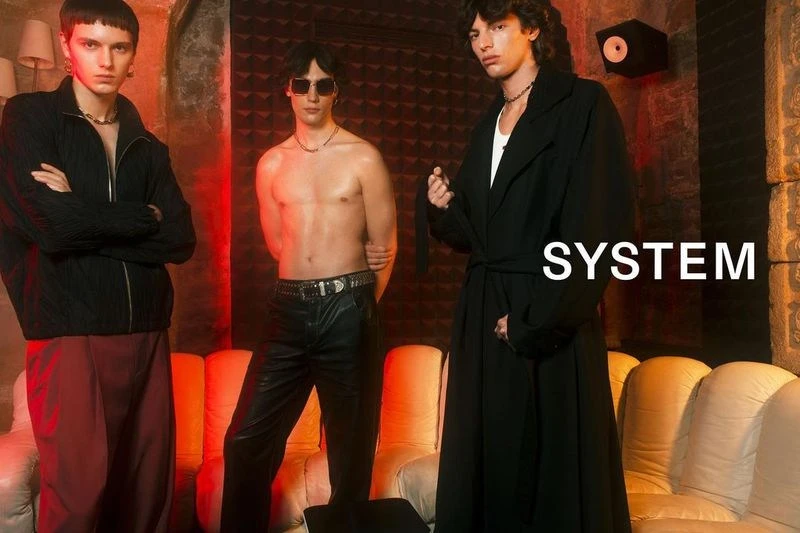 Three male fashion models pose in a warm-toned photo studio. One man is shirtless, another wears a black jacket and the third, a long black coat.
