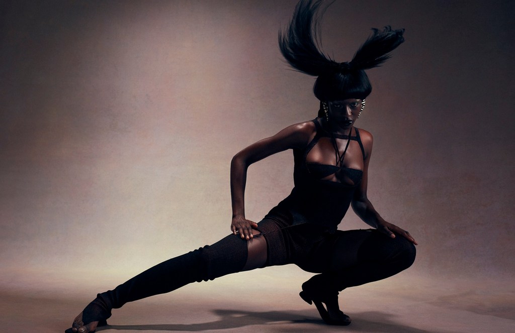 A striking, full-body shot of a Black woman in a unique, dark fashion outfit posing in a crouched position against a muted background.