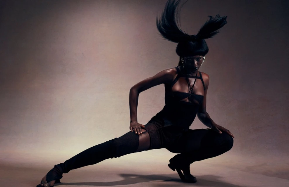 A striking, full-body shot of a Black woman in a unique, dark fashion outfit posing in a crouched position against a muted background.