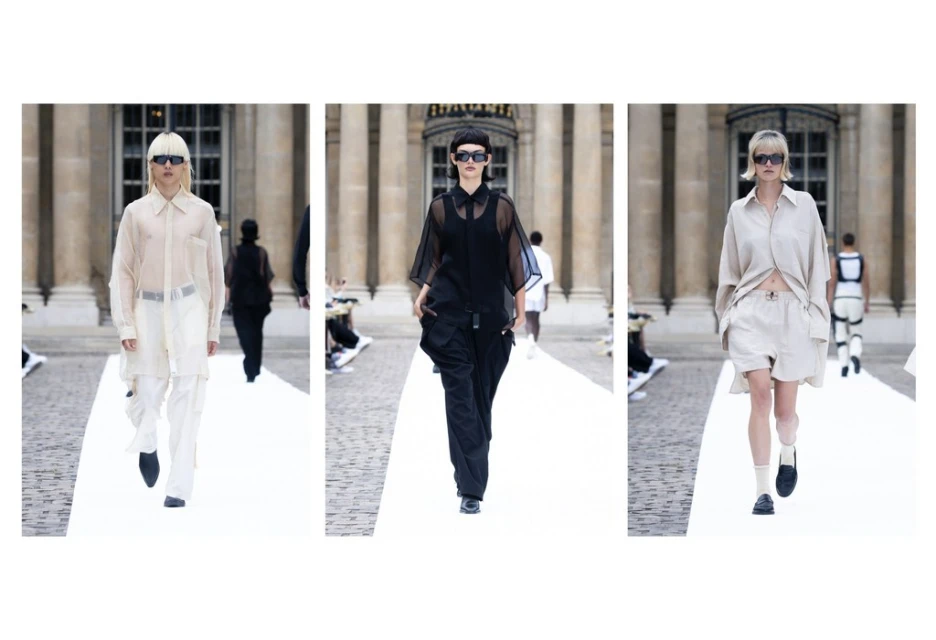 Fashion show featuring models walking on a white runway in front of an architectural building wearing stylish neutral-tone outfits and sunglasses.