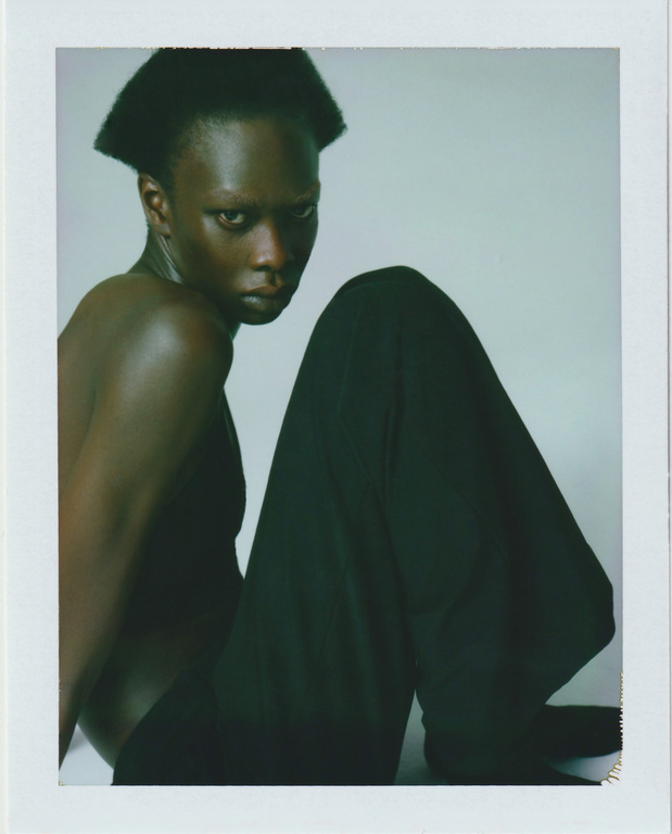 Portrait of a topless African female fashion model with black wide-leg pants, seated in a photo studio against a gradient gray background.