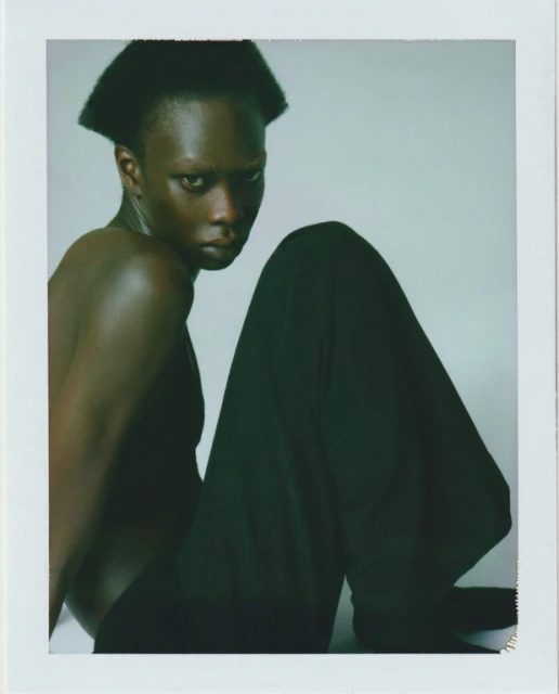 Portrait of a topless African female fashion model with black wide-leg pants, seated in a photo studio against a gradient gray background.