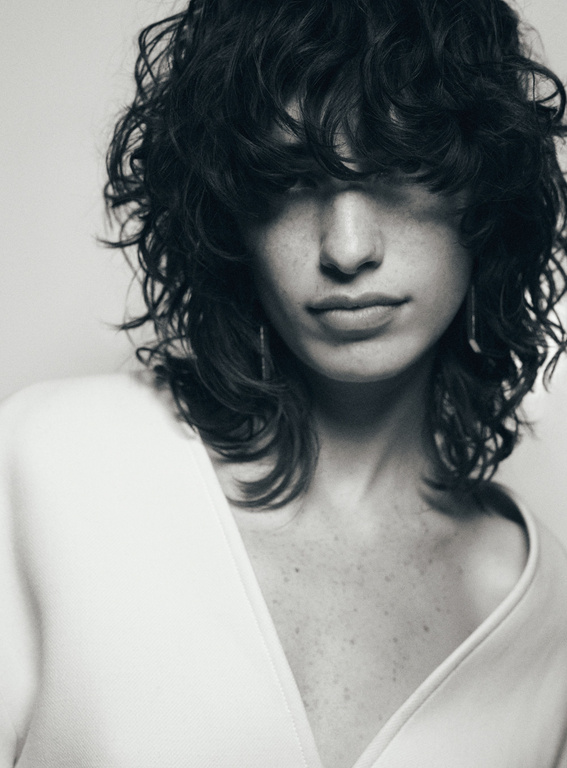 Monochrome portrait of a young fair-skinned woman with curly dark hair, wearing a light-colored V-neck top, against a bright background.