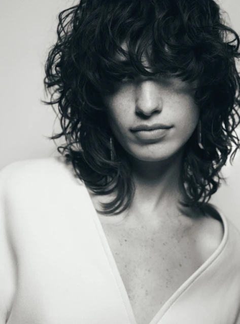 Monochrome portrait of a young fair-skinned woman with curly dark hair, wearing a light-colored V-neck top, against a bright background.