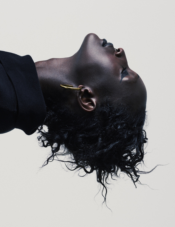 Dramatic studio portrait of a Black woman with dark curly hair and a gold earring, shot from a low angle with a white background.
